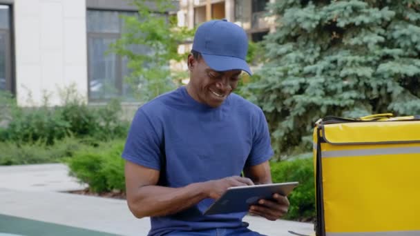 Sonriente hombre afro-americano mensajero entrega de alimentos sentado en el banco utiliza tableta — Vídeo de stock