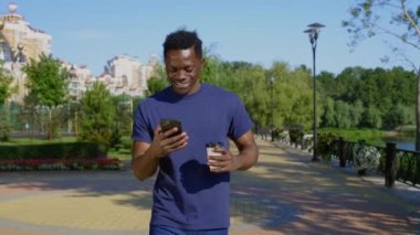 Gülümseyen Afro-Amerikalı adam sokakta yürürken cep telefonu bardağı ve kahve kullanıyor.