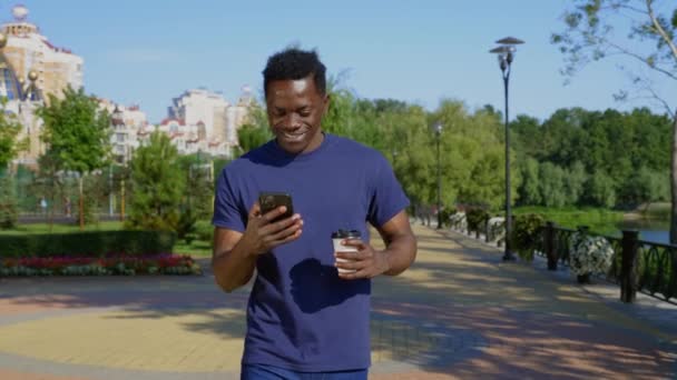 Sonriente hombre afroamericano camina calle utiliza teléfono móvil sosteniendo taza con café — Vídeos de Stock