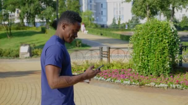 Sonriente hombre afroamericano camina calle utiliza teléfono móvil sosteniendo taza con café — Vídeos de Stock