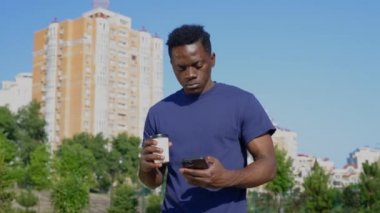 Afro-Amerikalı adam elinde kahve ya da çay olan beyaz bardakla cep telefonu kullanıyor.