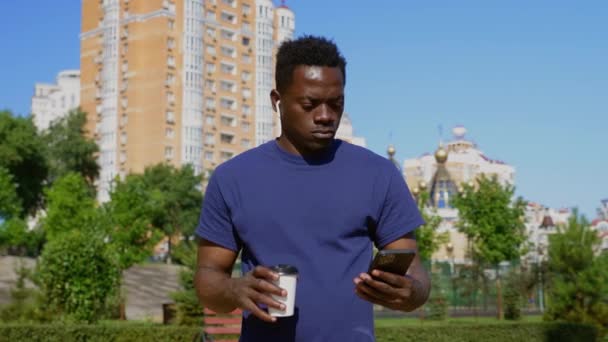 Porträtt Afrikansk man bär trådlösa hörlurar använder smartphone dricka kaffe — Stockvideo