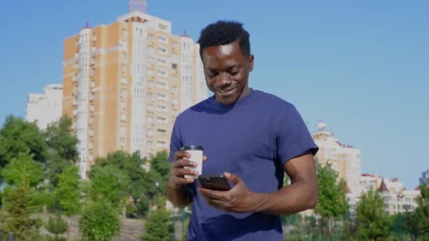 Sonriente hombre afroamericano camina calle utiliza teléfono móvil sosteniendo taza con café — Vídeos de Stock