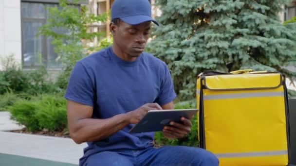 Zoom fotocamera in afro-americano uomo corriere consegna seduto su panca utilizza tablet — Video Stock