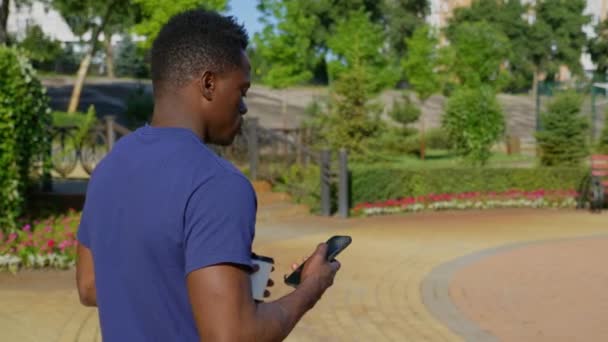 Hombre afroamericano utiliza teléfono móvil sosteniendo taza blanca con café o té en la mano — Vídeos de Stock
