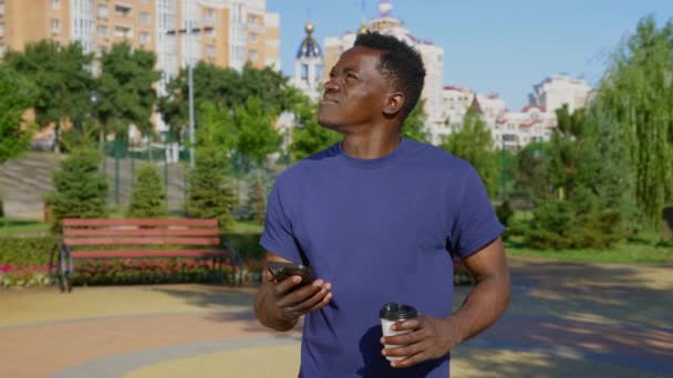 Homme afro-américain réfléchi debout dans le parc regarde autour, navigateur smartphone — Video