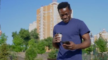 Gülümseyen Afro-Amerikalı adam sokakta yürürken cep telefonu bardağı ve kahve kullanıyor.