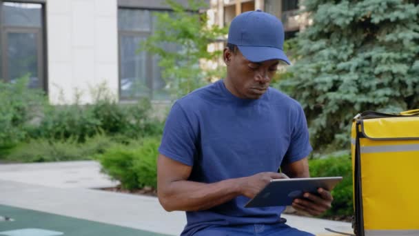 Consegna corriere uomo afro-americano seduto su panchina con zaino utilizza tablet — Video Stock