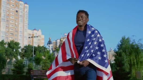 Un homme afro-américain souriant marche dans la rue avec le drapeau américain sur le dos en été — Video