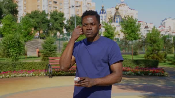 Retrato hombre adulto afroamericano de pie en el parque con auriculares inalámbricos — Vídeos de Stock