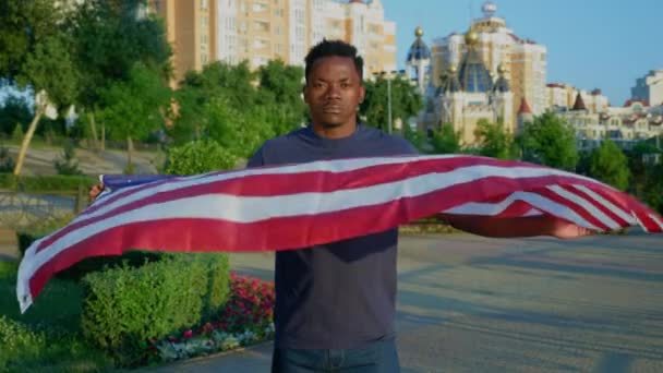 Портрет афро-американца, держащего американский флаг и смотрящего в камеру летом — стоковое видео