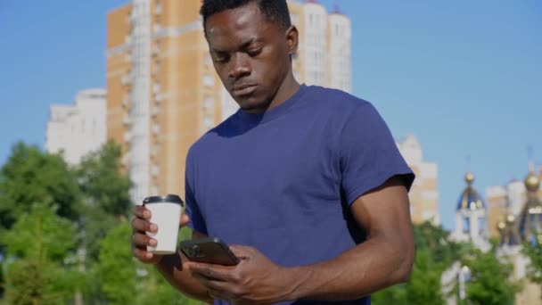 Hombre afroamericano utiliza teléfono móvil sosteniendo taza blanca con café o té en la mano — Vídeos de Stock