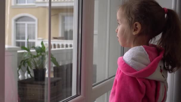 Dzieciak siedzi sam na parapecie i patrzy przez okno. Smutna dziewczynka podczas kwarantanny od wirusa korony wewnątrz. Słodkie dziecko w domu z powodu pandemii koronawirusowej. Bezpieczeństwo od COVID-19 — Wideo stockowe