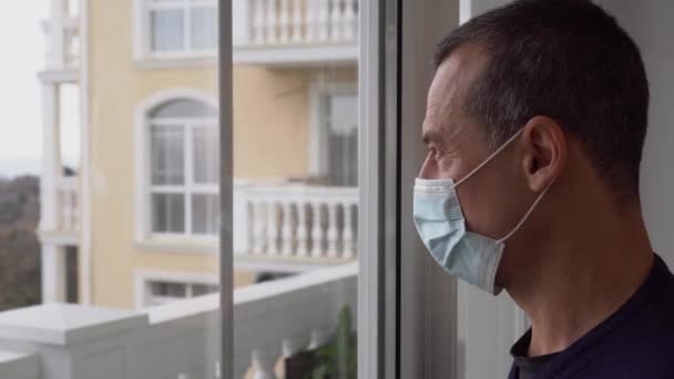 Zamykamy, zostajemy w domu. Samotny mężczyzna w średnim wieku w ochronnych maskach medycznych smutny patrzy przez okno na miasto. Izolacja domowa. Pandemia koronawirusa COVID-19 — Wideo stockowe