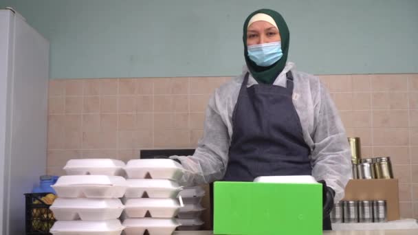 Voluntário de Apoio do Ramadã. Mulher muçulmana voluntária vestindo hijab, máscara e luvas. Cozinhar alimentos, embalagem de refeições quentes em lancheiras, distribuição para os necessitados — Vídeo de Stock