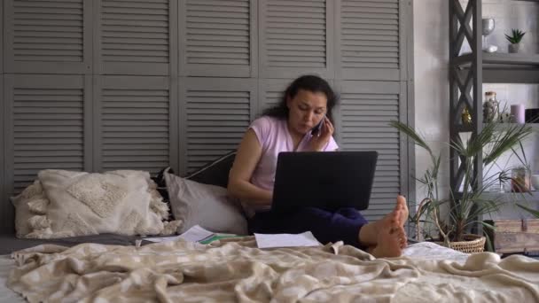 ラテンアメリカの女性は、ベッドの上の自宅で携帯電話でビジネス会話をしています。リモート、モバイルワーク、リモートワーク、柔軟な職場での作業 — ストック動画