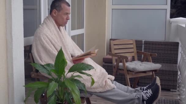 Un hombre de mediana edad se relaja en casa en el balcón, lee un libro, envuelto cómodamente en una cálida manta de invierno. Autoaislamiento bloqueo cuarentena estilo de vida durante la pandemia del Coronavirus — Vídeo de stock