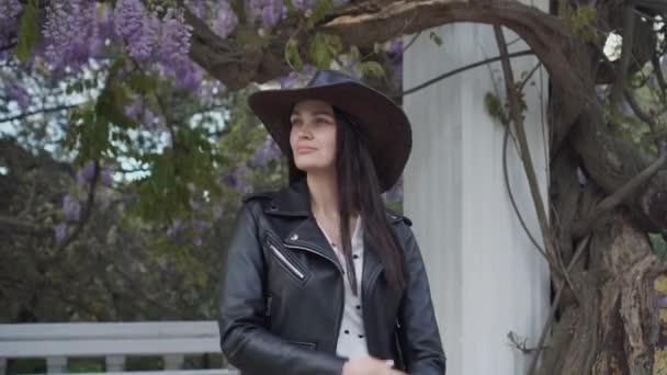 在这座城市的春天公园里，一个开着花的紫藤的背景上，有一个美丽的时髦的模型。一个年轻迷人的黑发女人，戴着帽子，穿着黑色皮夹克和漂亮的连衣裙 — 图库视频影像