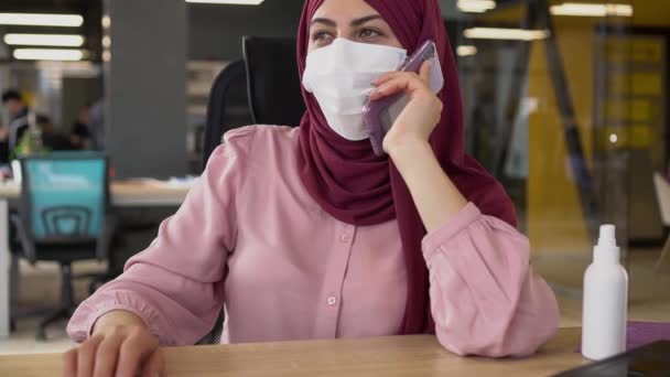 Modern muslimsk affärskvinna i hijab arbetar på kontoret och använder en smartphone under COVID-19 pandemin. Hälsosam arbetsplats för arbetstagare och kunder. Medicinsk mask och socialt avstånd — Stockvideo