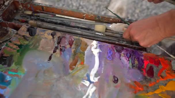 アーティストの手はブラシやツールを取ります。イーゼルに油絵具付きのパレット。画家は絵を描く。自然の中で絵画。プレイン・エア — ストック動画