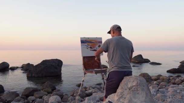 Sanatçı adam deniz manzaralı bir resim çiziyor. Şafakta taşlı deniz kıyısı. Yağlı boya boya doğada tuvale sürülmüş. Plein hava, manzara — Stok video