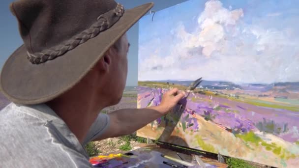 Ein männlicher Künstler zeichnet im Freien mit einem Palettenmesser ein Bild auf Leinwand. Plein air Ölgemälde. Ländliche Landschaft, blühende Lavendelfelder — Stockvideo