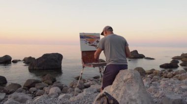 Doğada sanat terapisi. Sanatçı deniz ve kayalıkların resmini çiziyor, manzara, deniz manzarası ve yaz günbatımı. İyileşme ve Rehabilitasyon için hızlı yol. Fiziksel uzaklık, sosyal uzaklık