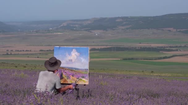 Plein lucht olieverf. Een mannelijke kunstenaar tekent een schilderij op doek buiten met een olieverf en penseel. Landelijk landschap, bloeiende lavendelvelden — Stockvideo