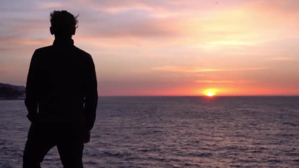 Сумний молодий самотній чоловік задумливо дивиться в океан. Прекрасний фон сходу сонця. Подорожі та самоізоляція, соціальне дистанціювання. Природа, море, пляж, ранок — стокове відео