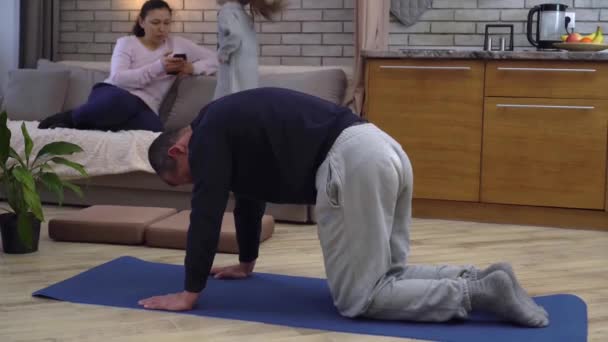 Un homme fait du yoga à la maison avec sa famille. Verrouillage du coronavirus et ordre de rester à la maison. Covid-19 et mode de vie sain du corps concept — Video