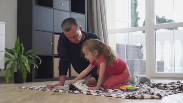 Χαρούμενη Ημέρα Πατέρων. Ένας πατέρας διαβάζει ένα βιβλίο στη μικρή του κόρη στο σπίτι, κάθεται στο πάτωμα. Covid-19 και Coronavirus κλείδωμα και διαμονή-στο-σπίτι παραγγελία. Πατέρας και παιδιά σε καραντίνα έχουν αστείο χρόνο — Αρχείο Βίντεο