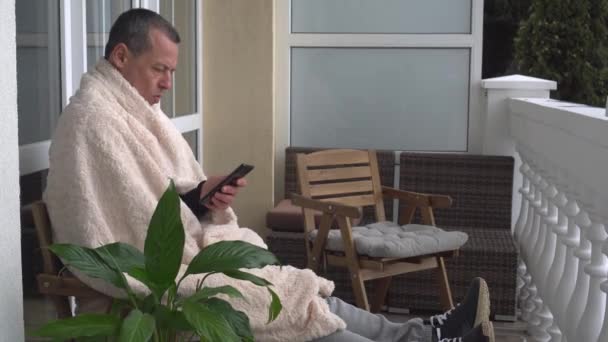 Un homme d'âge moyen se détend à l'automne à la maison sur le balcon, lit un livre, enveloppé confortablement dans une couverture d'hiver chaude. Auto-isolement mode de vie en quarantaine pendant la pandémie de coronavirus — Video