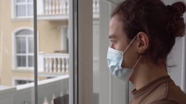 COVID-19 Coronavirus kilitlenmesi. Tıbbi maskeli üzgün genç adam pencereden şehre bakıyor. Kilitlenme ve ekonomik kriz. Karantina, kendini izole etme, evde kal.