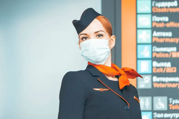 Beautiful Red Head Flight Attendant Wearing Protecting Mask and Stay Near Departure And Arrival Board. Covid-19 Outbreak Travel Restrictions. Flight Cancellation.