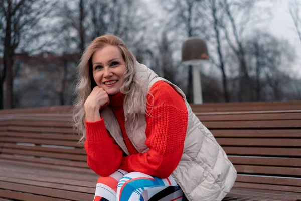 Зрелая блондинка улыбается и сидит на скамейке в парке после упражнений. Активный образ жизни пожилых людей — стоковое фото
