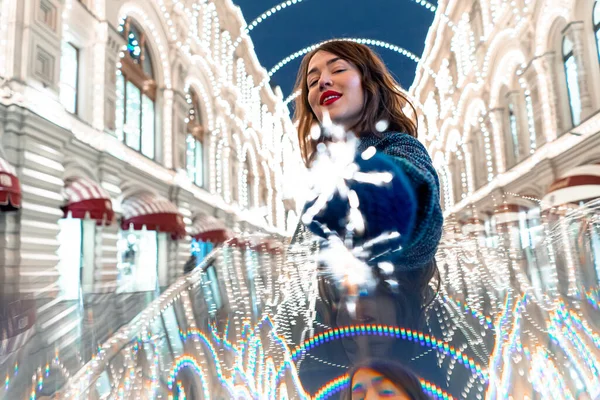 Allegro giovane donna che tiene Sparkler e sorridente vicino decorato con luci di Natale edificio. Festeggiamo Capodanno 2021 con la luce del Bengala — Foto Stock