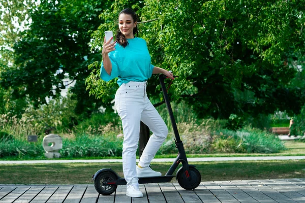 Junge Schöne Frau in weißen Jeans, hellblauem T-Shirt und weißen Turnschuhen in der Nähe von schwarzem Elektroroller im Gespräch via Videoanruf im Park — Stockfoto