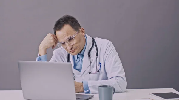 Tired doctor in medical uniform working on computer takes a sip of coffee