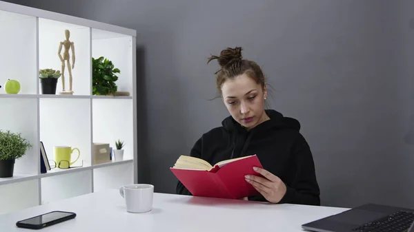 Hermosa chica con capucha negra es interesante leer libro en casa y sonreír. — Foto de Stock