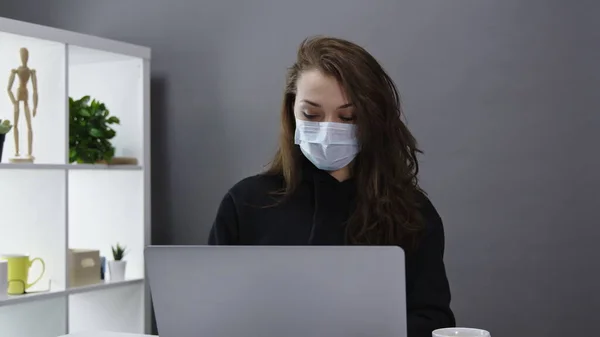 Linda mujer de negocios en máscaras médicas trabaja en el ordenador portátil en la oficina en casa, trabajo remoto — Foto de Stock