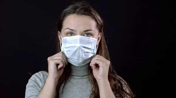 Mulher bonita coloca máscara médica. Fundo preto. Covid-19 pandémico global — Fotografia de Stock