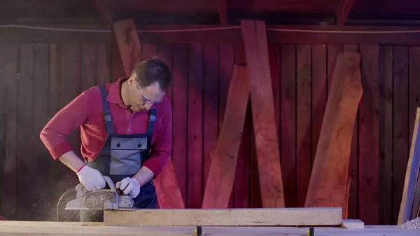 Male carpenter cleans surface of wooden beam using electric planer in workshop — Stock Photo, Image