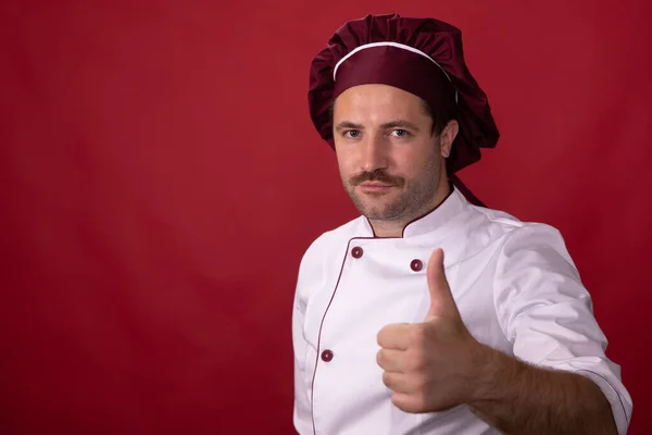 Unshaven guapo macho chef muestra el pulgar hacia arriba mirando la cámara en rojo telón de fondo —  Fotos de Stock