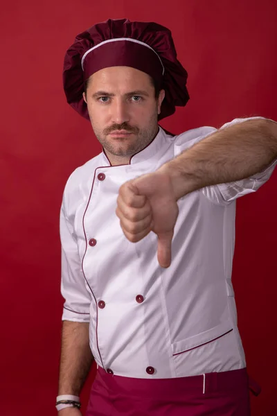 Unshaven guapo macho chef muestra el pulgar hacia abajo mirando a la cámara en rojo telón de fondo —  Fotos de Stock