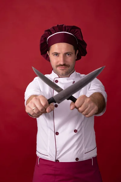 Unshaven guapo chef posando cuchillos cruzados en las manos sobre fondo rojo estudio —  Fotos de Stock