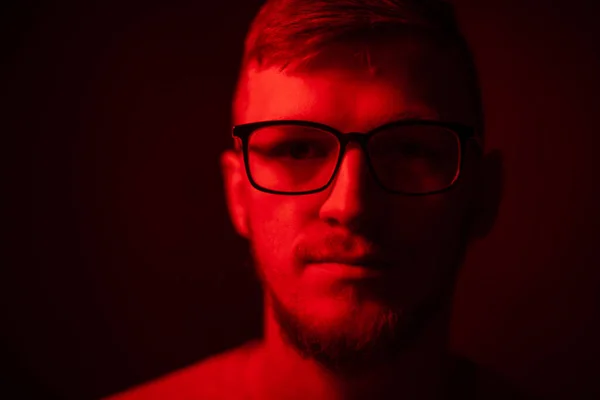 Low key portrait of bearded young man in glasses looking at camera in red light — Stock Photo, Image