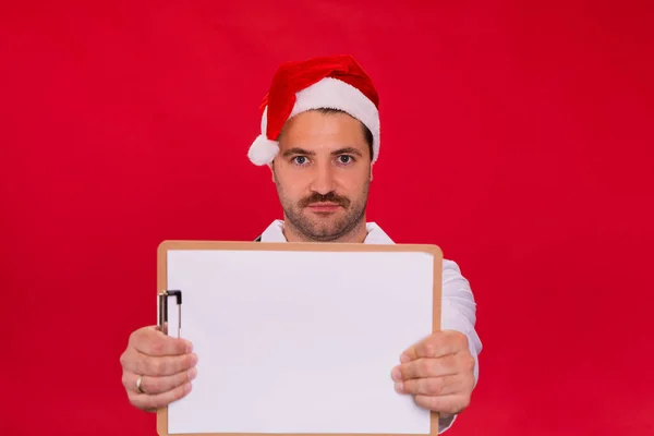 Médico masculino sin afeitar en sombrero de Santa Claus sosteniendo portapapeles en blanco maqueta — Foto de Stock