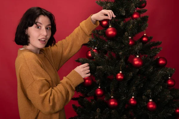 Sonriente vestida casualmente morena chica cuelga pelotas de juguete en árbol de Navidad en rojo — Foto de Stock