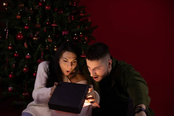 Pareja romántica sorprendida de abrir regalo mágico de Navidad sobre fondo rojo oscuro — Foto de Stock