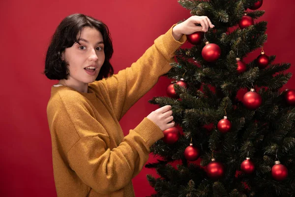 Joven mujer excitada en jersey de invierno decorando árbol de Navidad sobre fondo rojo —  Fotos de Stock